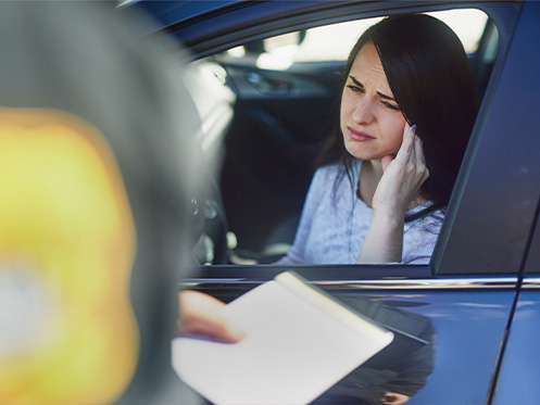 A imagem mostra uma mulher dentro de seu carro com os dedos pressionados sobre sua cabeça, olhando um policial à entregar uma multa