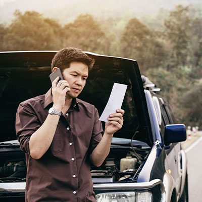 a imagem ilustra um homem encostado no capô do carro (que está aberto) segurando um papel e telefonando para alguém