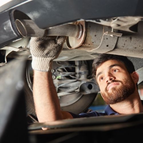 A imagem mostra um homem trabalhando embaixo de um carro que está levantado por um elevador automotivo. O homem usa luvas e uma camisa azul escura com gola.