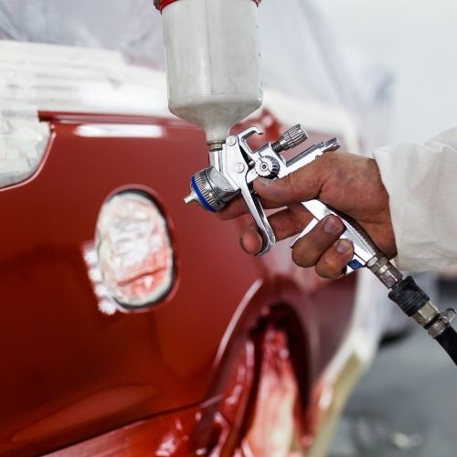 A imagem mostra um profissional de pintura aplicando uma tinta vermelha em um carro vermelho. A tinta é aplicada com uma pistola de pintura.