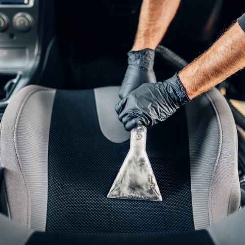 A imagem mostra duas mãos usando luvar pretas segurando a ponta de extrator enquanto passa na parte de trás do banco de um carro.