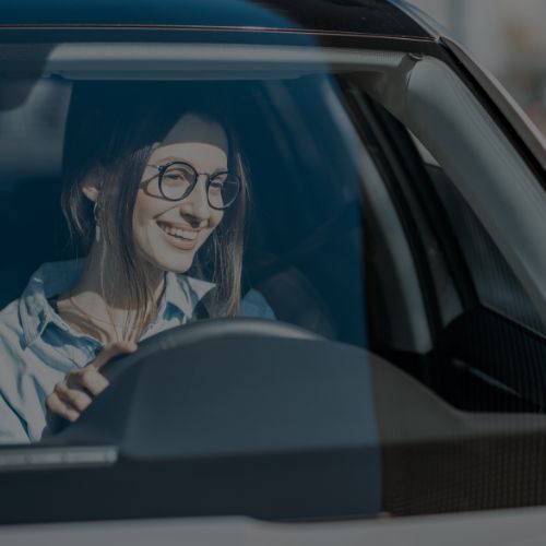 A imagem mostra uma mulher vista de fora do carro, pelo para-brisa, dirigindo o carro