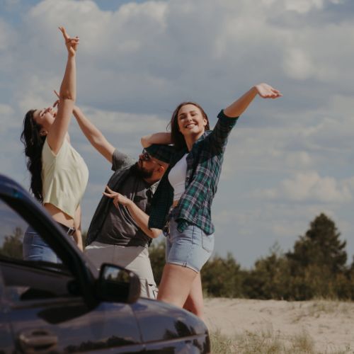A imagem mostra três jovens se divertindo ao ar livre em um dia ensolarado. Eles estão ao lado de um carro preto, parcialmente visível no canto esquerdo da imagem. Duas das jovens estão levantando os braços, sorrindo e parecendo animadas, enquanto o jovem no meio faz um gesto expressivo com as mãos.