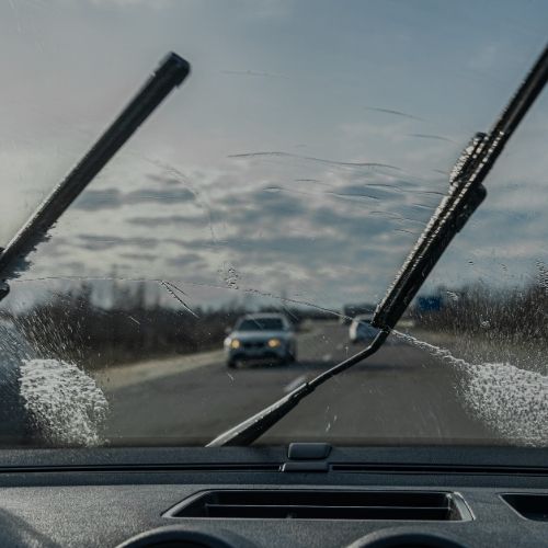 Imagem vista do interior de um carro, mostrando limpadores de para-brisa em ação em um dia nublado, com carros ao fundo na estrada, destacando segurança na condução.