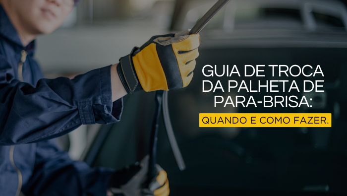 Pessoa com luvas amarelas e pretas segurando uma palheta de para-brisa durante sua troca, com o vidro do carro desfocado ao fundo, em um ambiente de oficina.